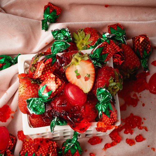 Mismatched strawberry candy stud earrings by Yellow Owl Workshop resting on a sliced strawberry among fresh berries, red jelly candies, and retro strawberry-wrapped sweets. Gold-tone enamel earrings inspired by old-fashioned strawberry bonbons.