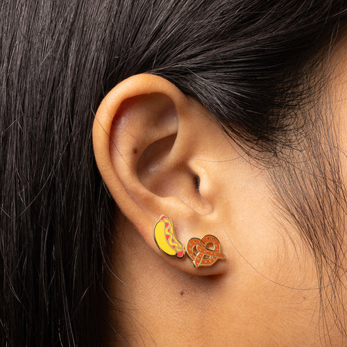 Hot dog and pretzel mismatched cloisonné stud earrings worn on ear by Yellow Owl Workshop