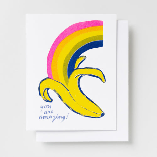 Risograph greeting card with an illustrated banana peel with a bold rainbow bursting from it in pink, orange, yellow, and navy on a white background, text reads You Are Amazing in handwritten blue lettering