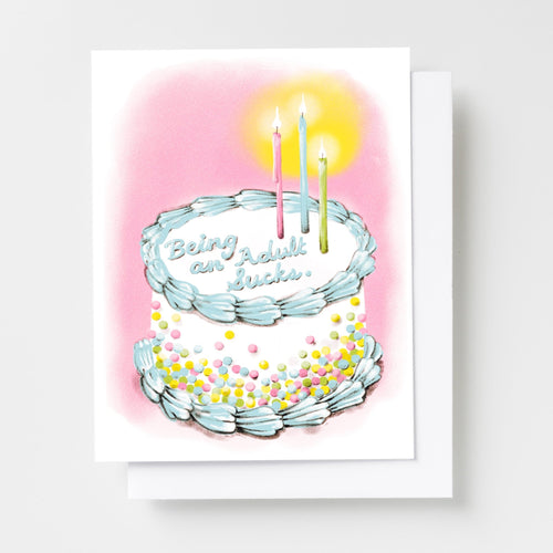 Risograph greeting card with an illustrated two-tier birthday cake with sprinkles and candles on a pink background, cake reads Being an Adult Sucks in cursive frosting lettering