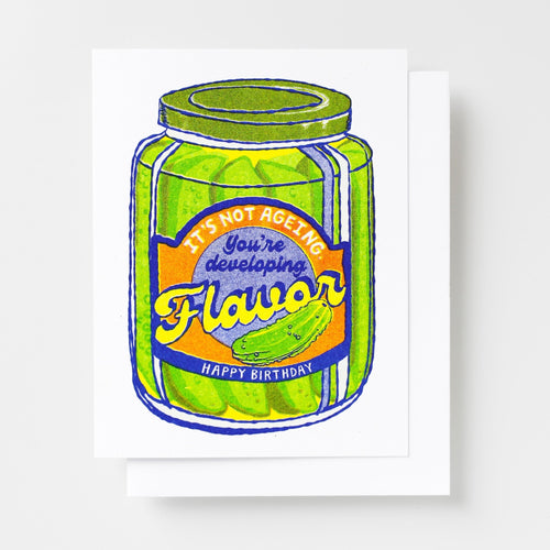 Risograph greeting card with an illustrated pickle jar with a label reading It's Not Ageing You're Developing Flavor Happy Birthday in green, yellow, and blue