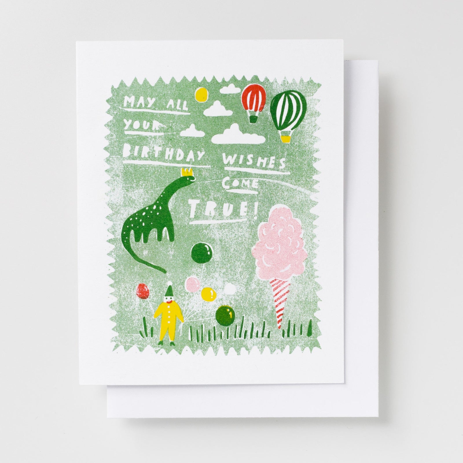 Risograph greeting card with a crowned green dinosaur, clown, cotton candy, and hot air balloons on a sage green background, text reads May All Your Birthday Wishes Come True in white lettering