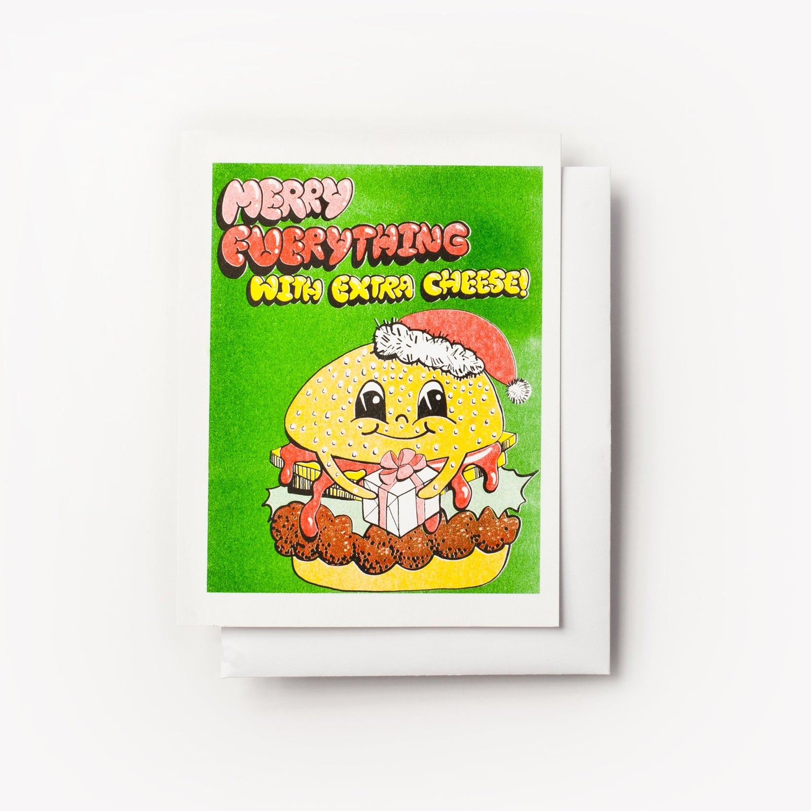 Risograph greeting card with an illustrated burger character wearing a Santa hat and holding a gift with melted cheese dripping on a bright green background, text reads Merry Everything with Extra Cheese in bold multicolor lettering