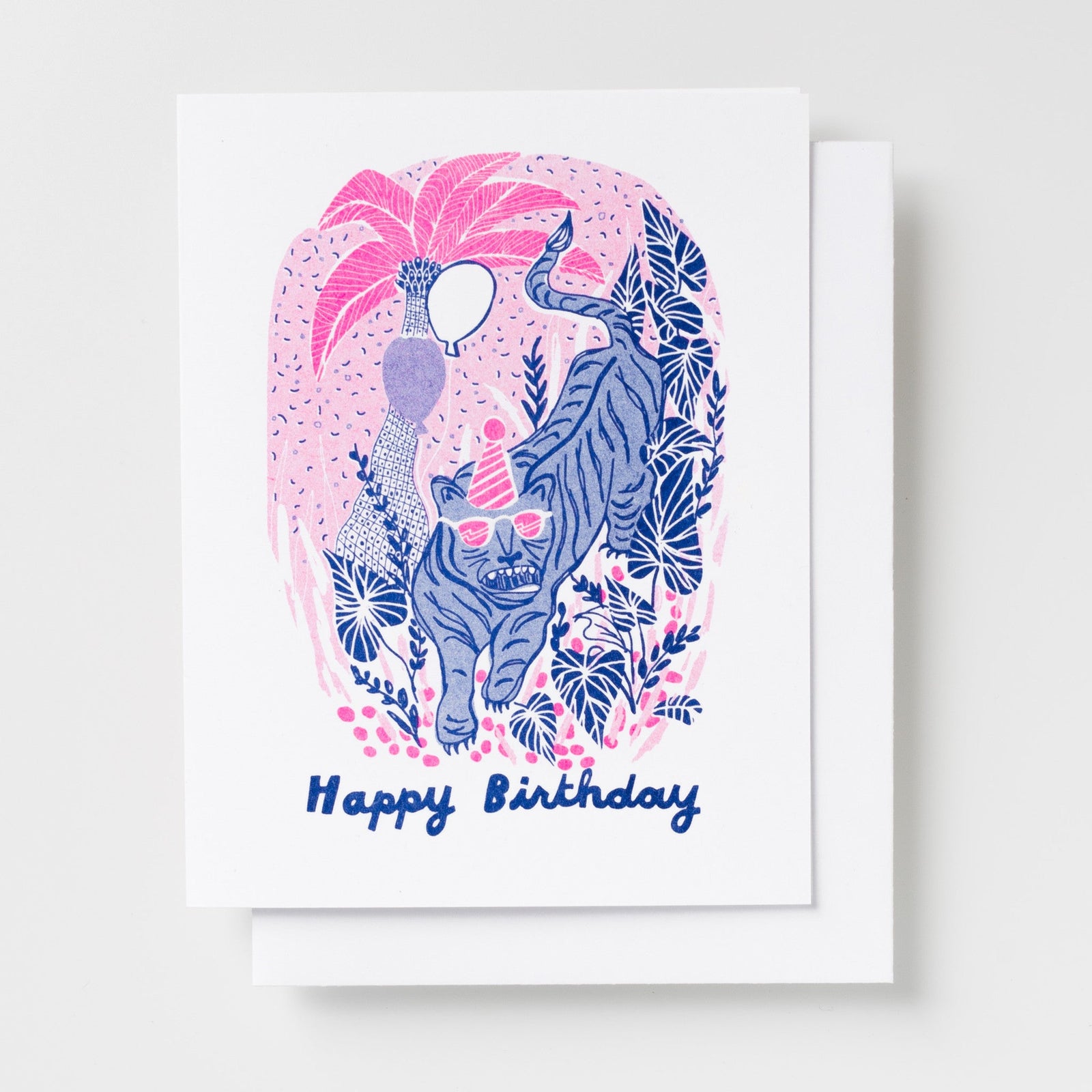 Risograph greeting card illustrated with a blue tiger wearing a party hat among tropical foliage and flowers in pink and navy, text reads Happy Birthday in cursive script