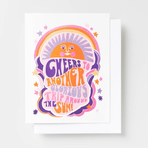 Risograph greeting card with a retro illustrated sun, rainbow, and stars in purple, orange, and pink, text reads Cheers to Another Glorious Trip Around the Sun in curved lettering